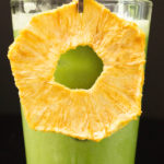 A bright green iced matcha drink garnished with a dried pineapple ring in a tall glass on a black background