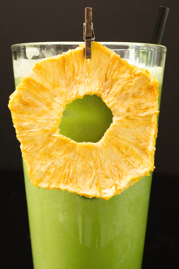 A bright green iced matcha drink garnished with a dried pineapple ring in a tall glass on a black background.