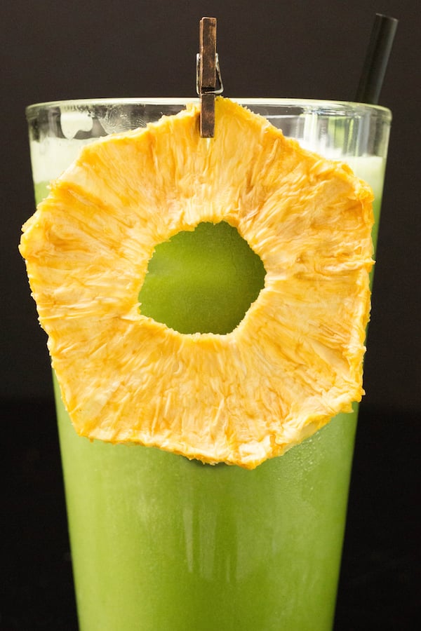 A bright green iced matcha drink garnished with a dried pineapple ring in a tall glass on a black background