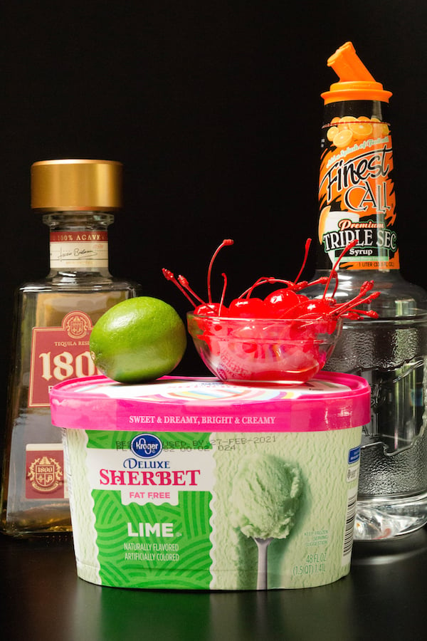 Ingredients for lime sherbet margaritas (lime sherbet, tequila, triple sec, and maraschino cherries) on a black background.
