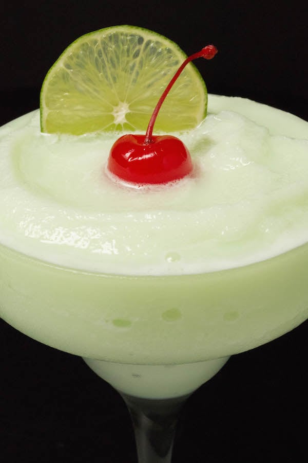 Close up of a lime sherbet margarita garnished with a cherry and lime slice on a black background.