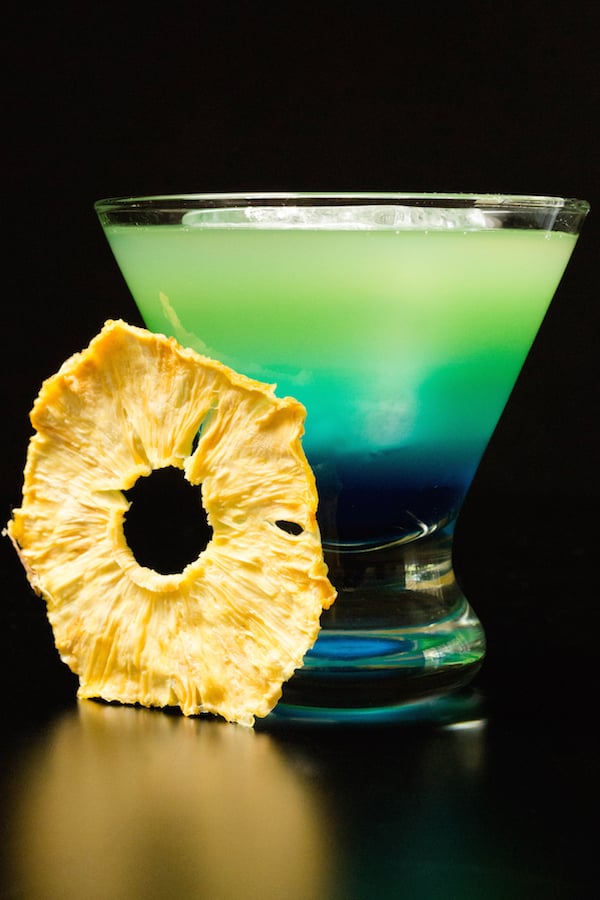 A layered blue to green Quarantiki cocktail in a glass on a blackbackground.