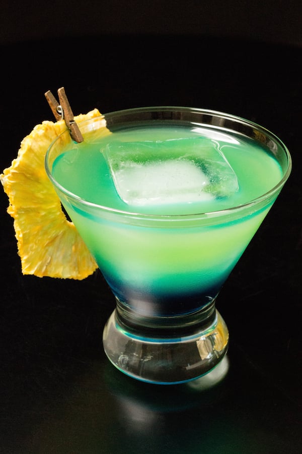 A blue to green layered cocktail garnished with a pineapple slice in a glass on a black background.