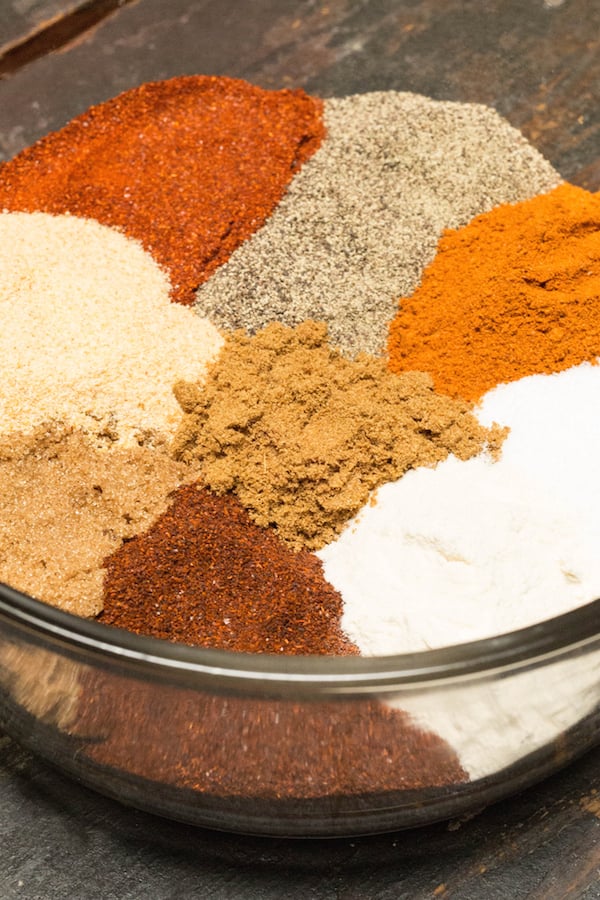 Multi-colored spices for BBQ spice rub arranged in piles in a large glass bowl.