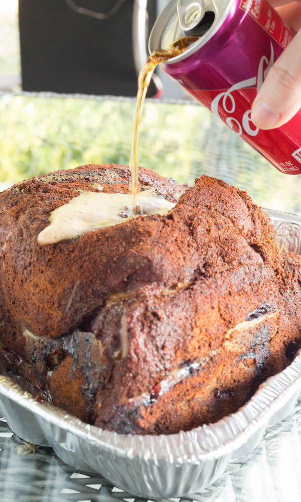 Cherry coke from a can being poured over the top of a cook pork should that is sitting in a disposable aluminum pan