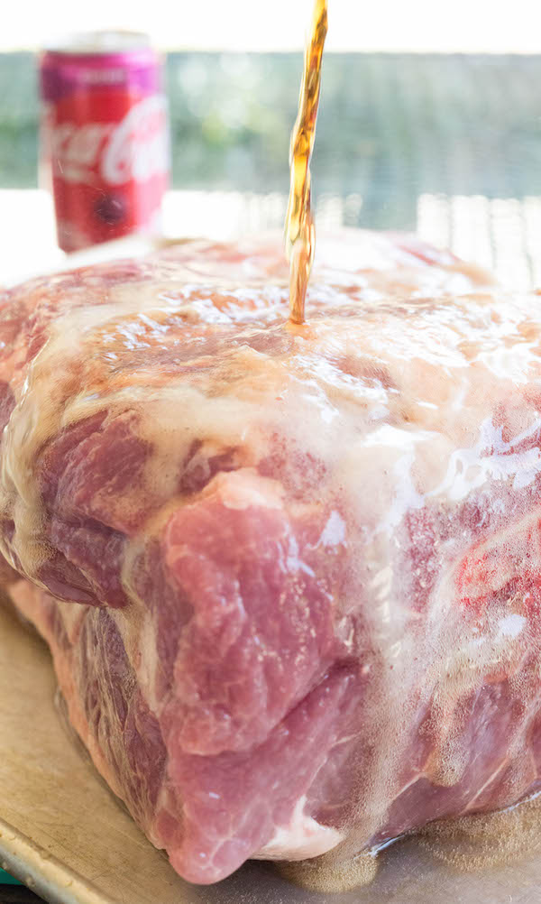 Cherry coke being poured over a raw pork shoulder on a baking sheet