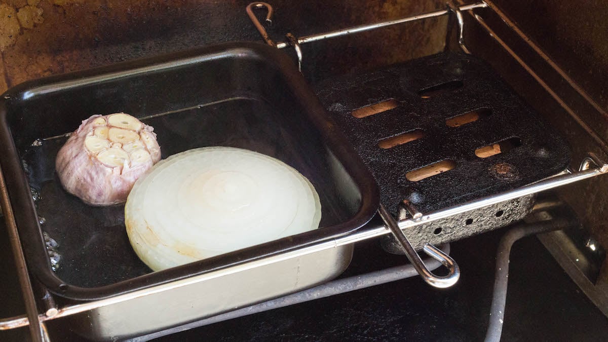 A meat smoker's waterpan filled with water, a garlic bulb, and half of an onion