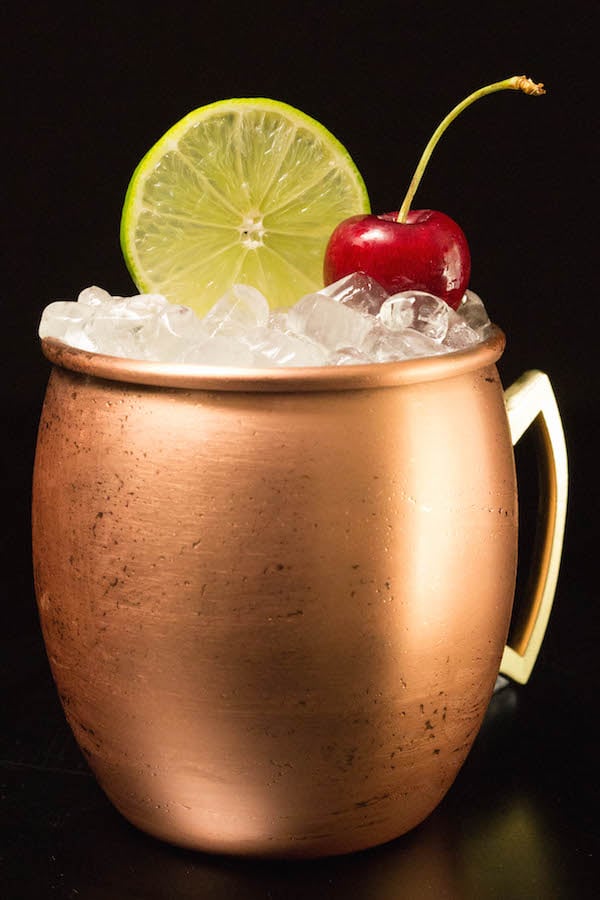 A copper mug filled with cherry lime moscow mule cocktail sits on a black background