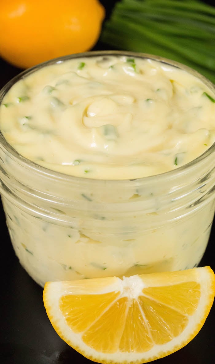 A small jar of lemon chive mayo sits on a black table with chives and a lemon in the background