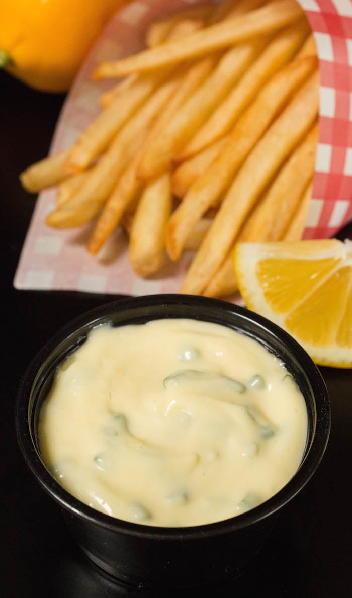 A small container of lemon chive mayo with french fries and a sliced lemon in the background