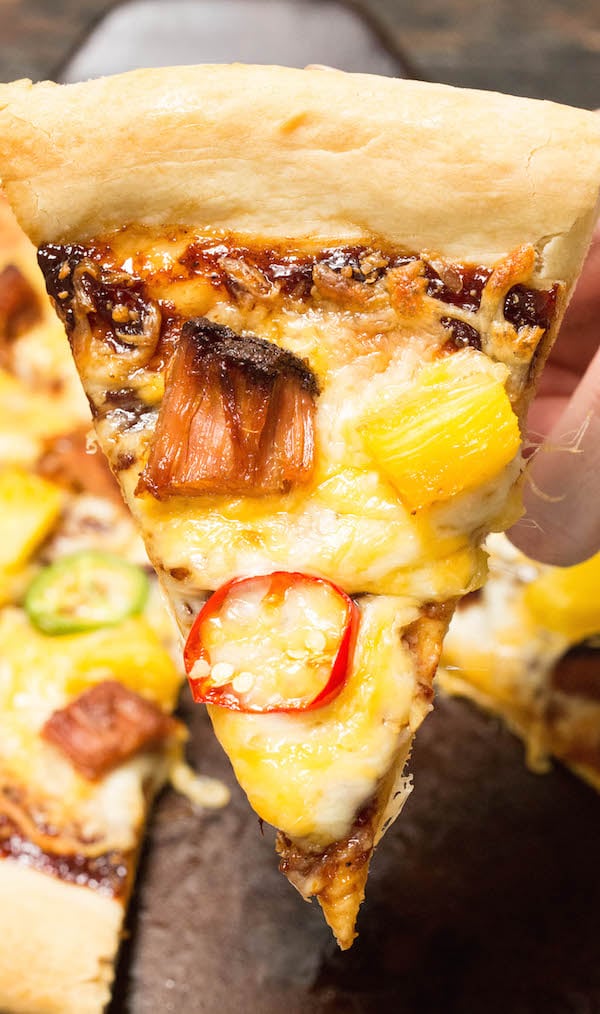 A slice of pork & pineapple bbq pizza being held up by a white hand.