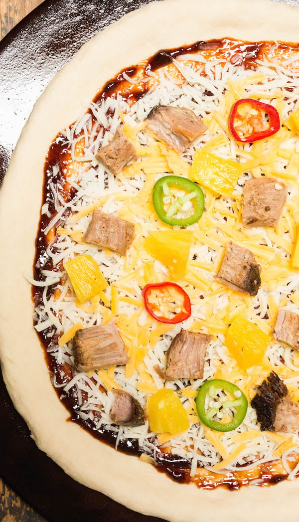 Raw pizza dough topped with BBQ sauce, cheese, jalapenos, pineapples, and smoked pulled pork on a pizza stone.
