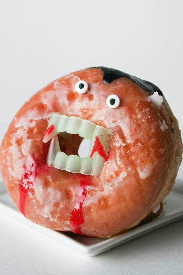 A glazed donut stands up vertically. Fake plastic vampire teeth are in the hole and candy eyes are on the top to make it look like a vampire. 