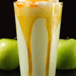 A glass of disney's caramel apple smoothie on a balck background with two green apples by it's side