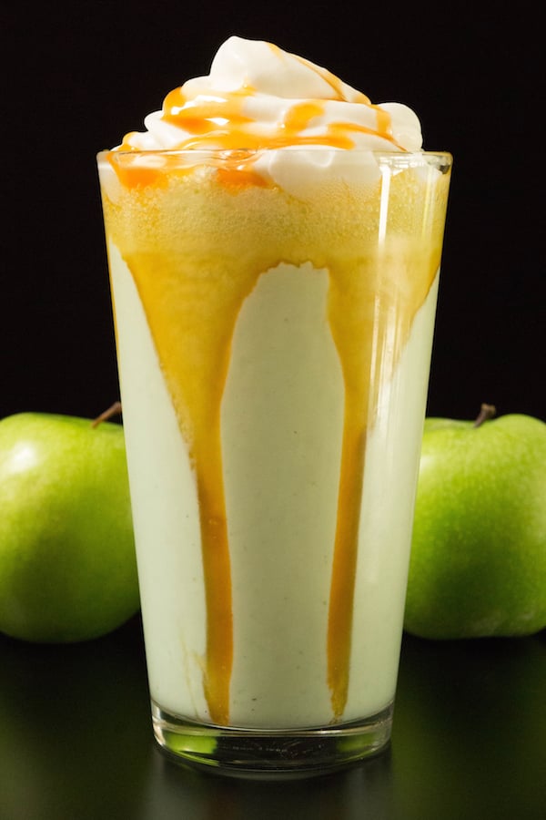 A glass of disney's caramel apple smoothie on a balck background with two green apples by it's side