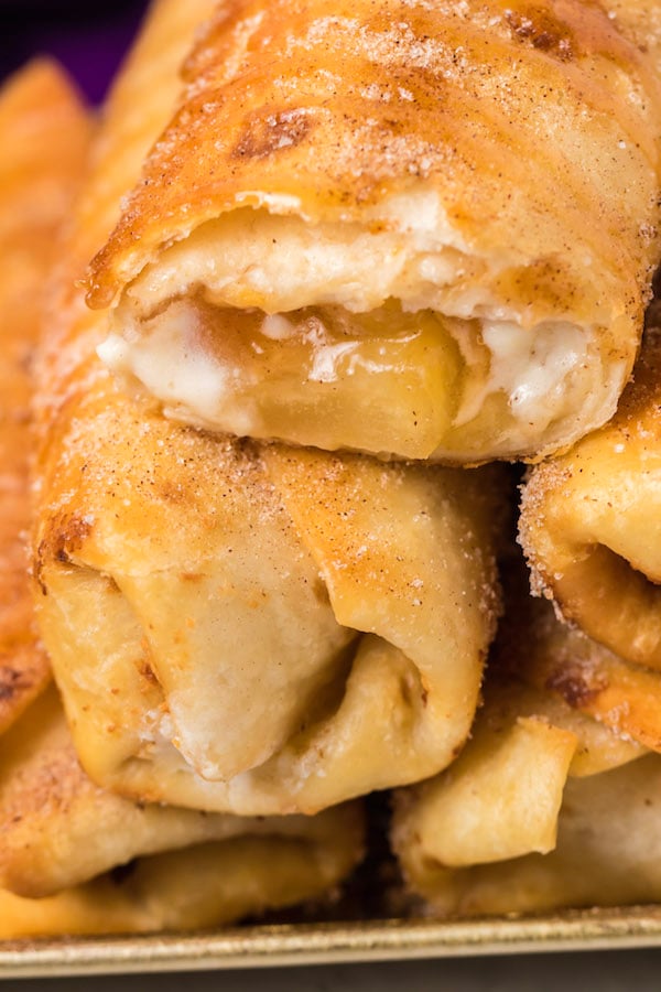 Close up of a stack of caramel apple cheesecake chimichangas. The on top is cut open to reveal the apple cheesecake filling inside.