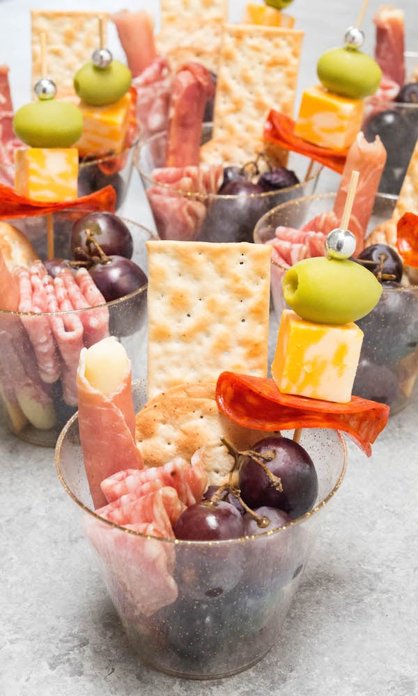 Four plastic cups filled with meat, cheese, fruit, and crackers on a bright background. 