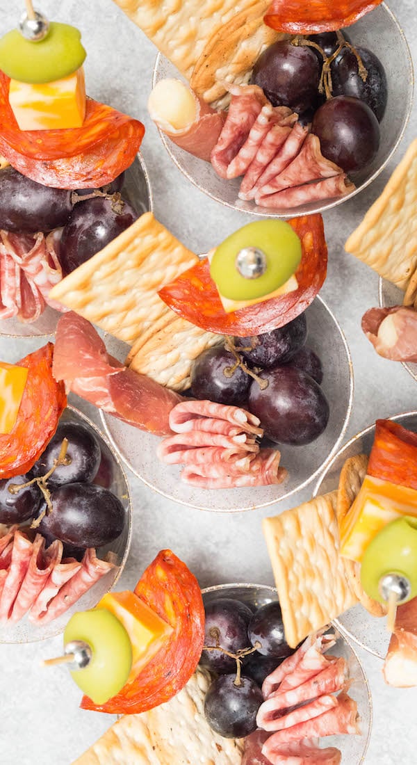 Overhead shot of plastic cups filled with meat, cheese, and grapes on a light background.