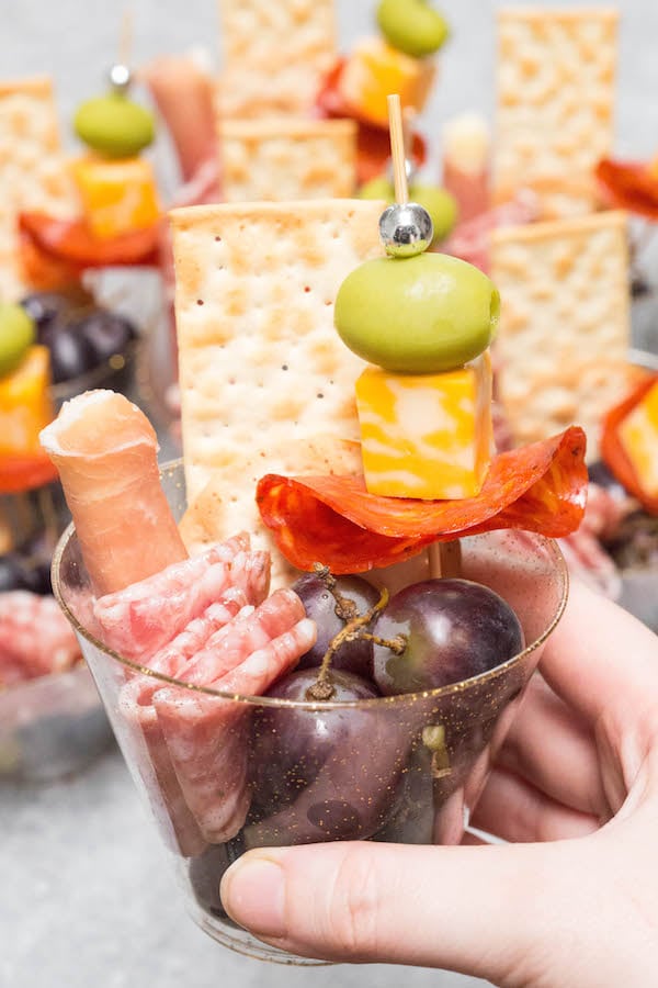 Close up of a hand holding a plastic charcuterie cup filled with meat, cheese, and grapes.