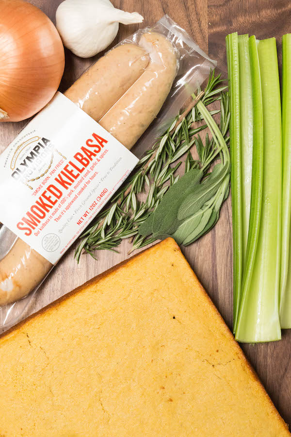 The ingredients for cornbread & sausage stuffing lie on a wood cutting board. A package of smoked kielbasa, rosemary, sage, celery, yellow onion, garlic, cornbread