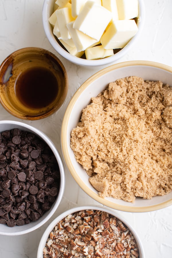 Overhead shot of 5 white bowls, each holding an ingredient for butter toffee (butter, vanilla, brown sugar, pecans, chocolate chips)