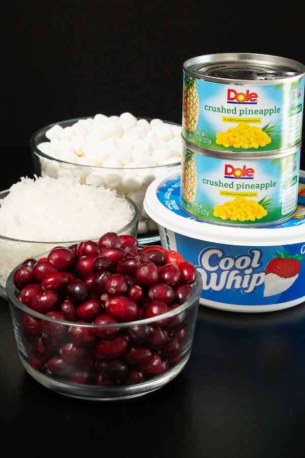 A tub of cool whip, two cans of crushed pineapple, a bowl of fresh cranberries, coconut, and mini marshmallows sit on a black background to show the ingredients needed for cranberry fluff