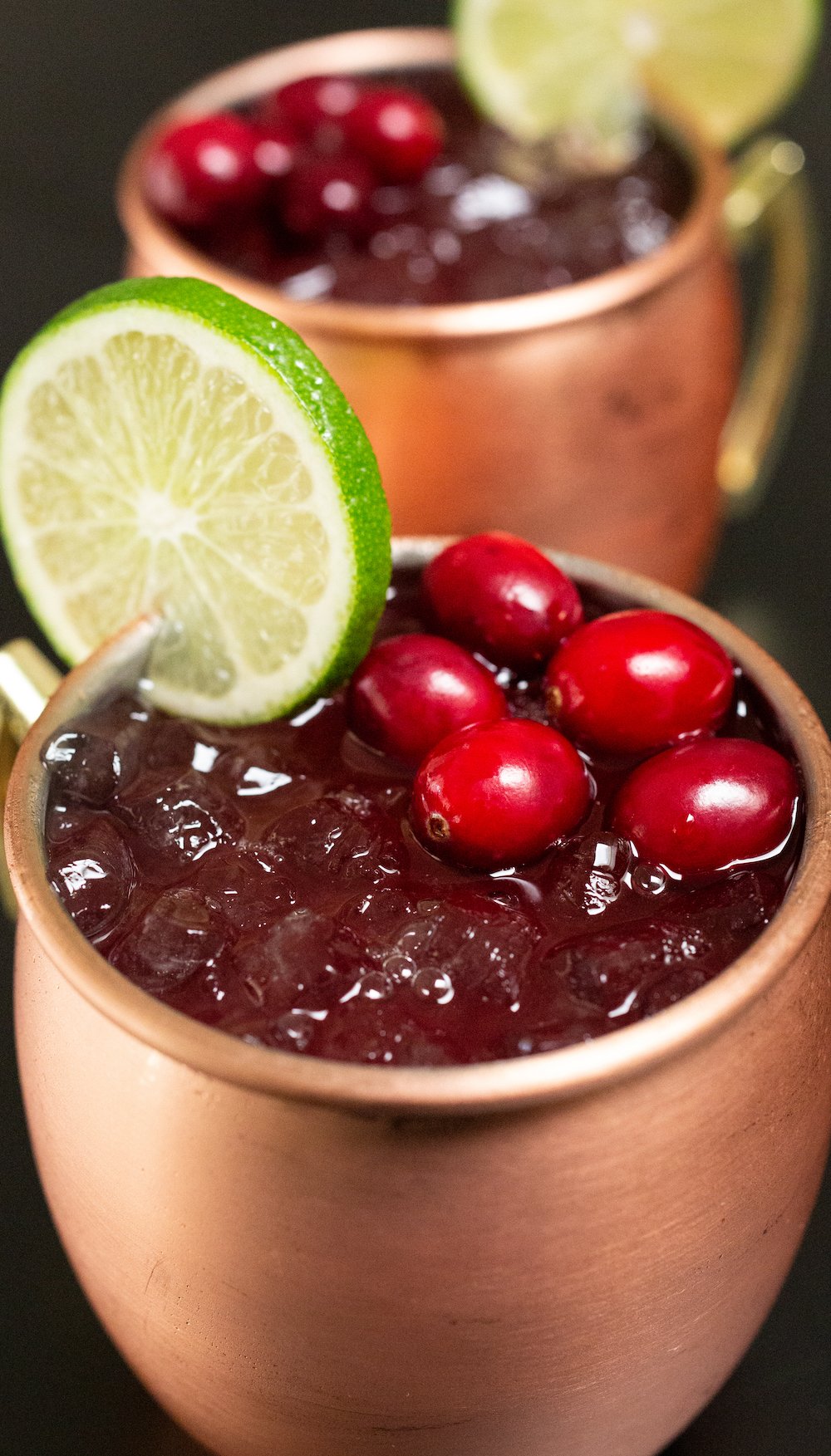 Two copper mules filled with cranberry moscow mules sit on a black background. They are each garnished with a lime wheel and fresh cranberries.