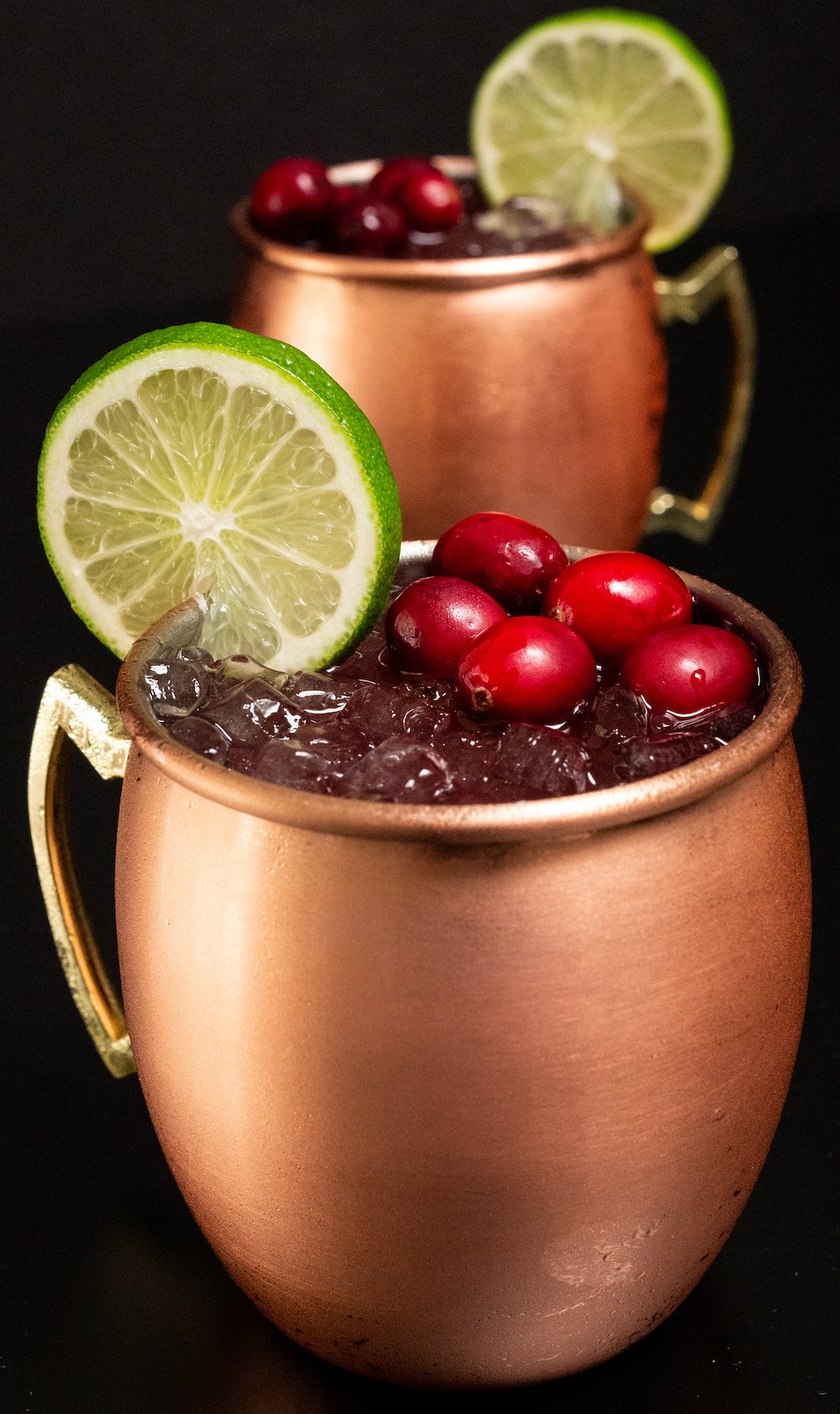 Wide shot of two cranberry moscow mules in copper mugs sitting on a black background.