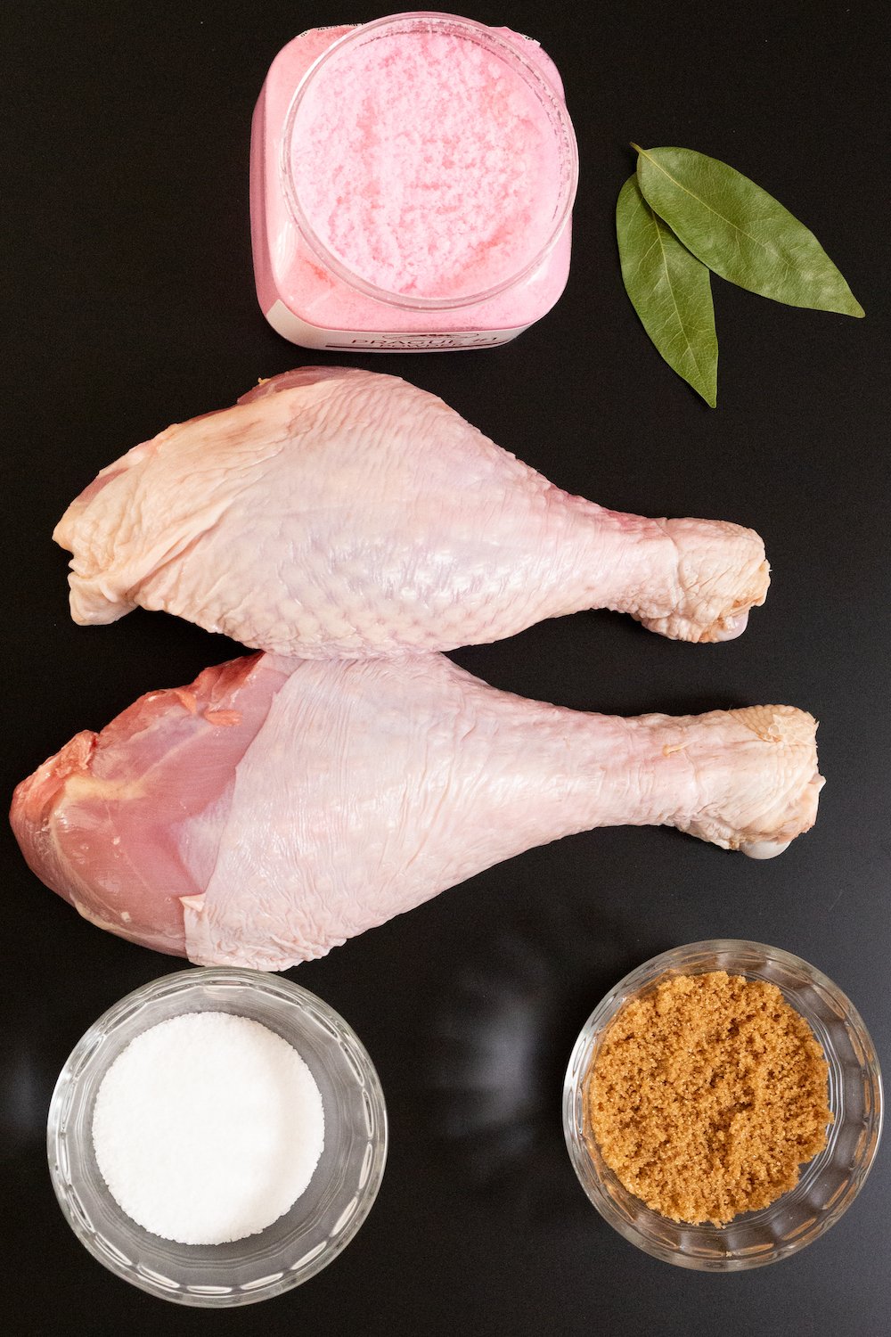 Overhead shot of a container of prague powder, two turkey legs, salt, brown sugar, and bay leaves 