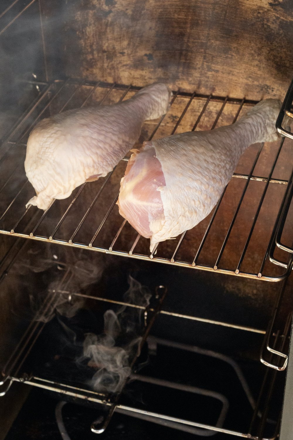 Two raw turkey legs sit on the middle rack of an electric smoker while smoke billows up