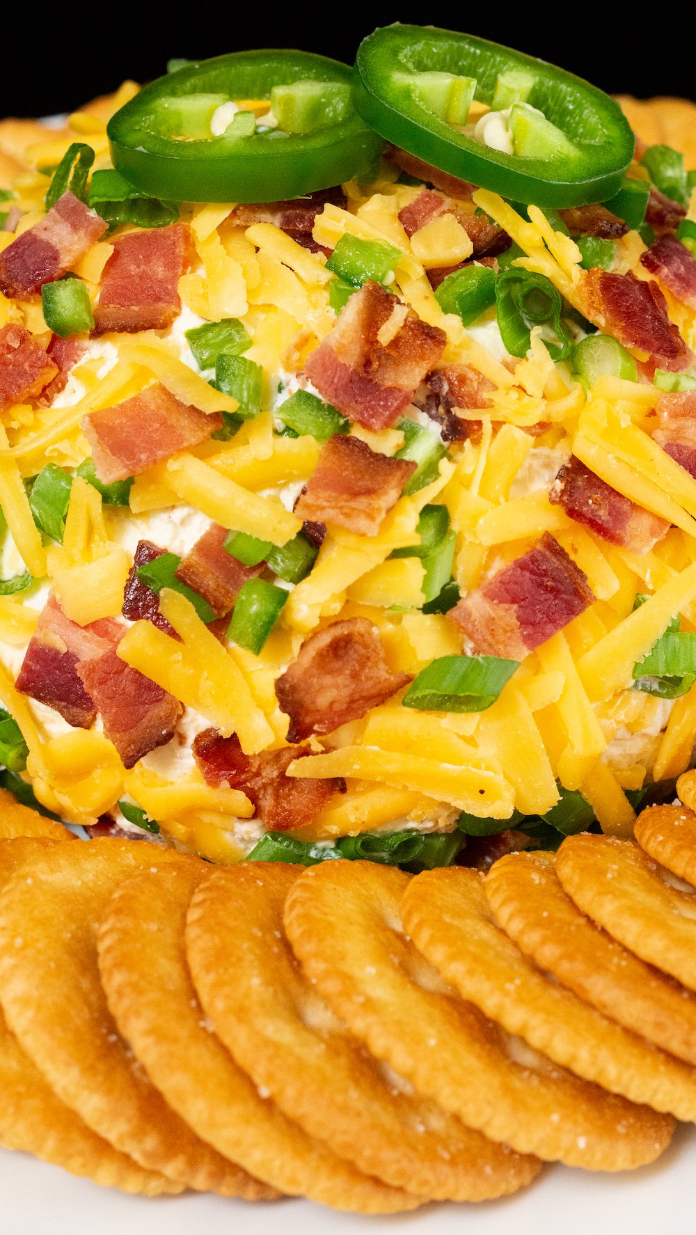 A cheese ball covered in shredded cheddar cheese, chopped, bacon, and chives, sits on the middle of a white plate surrounded by ritz crackers