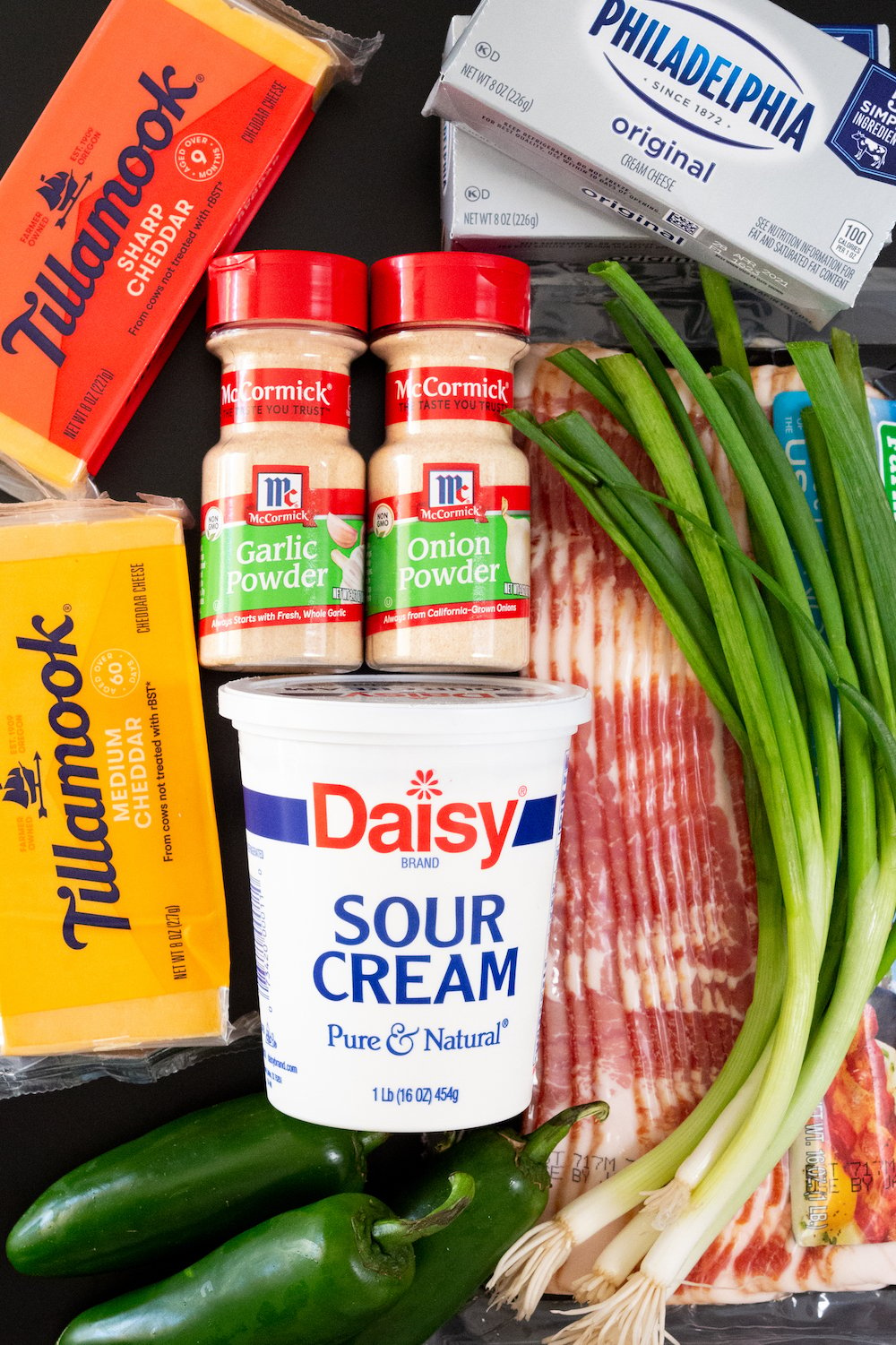 All the ingredients for a jalapeno popper cheese ball lay on a black table (2 blocks of cheddar cheese, cream cheese, onion powder, garlic powder, bacon, sour cream, green onions, and three fresh jalapenos).