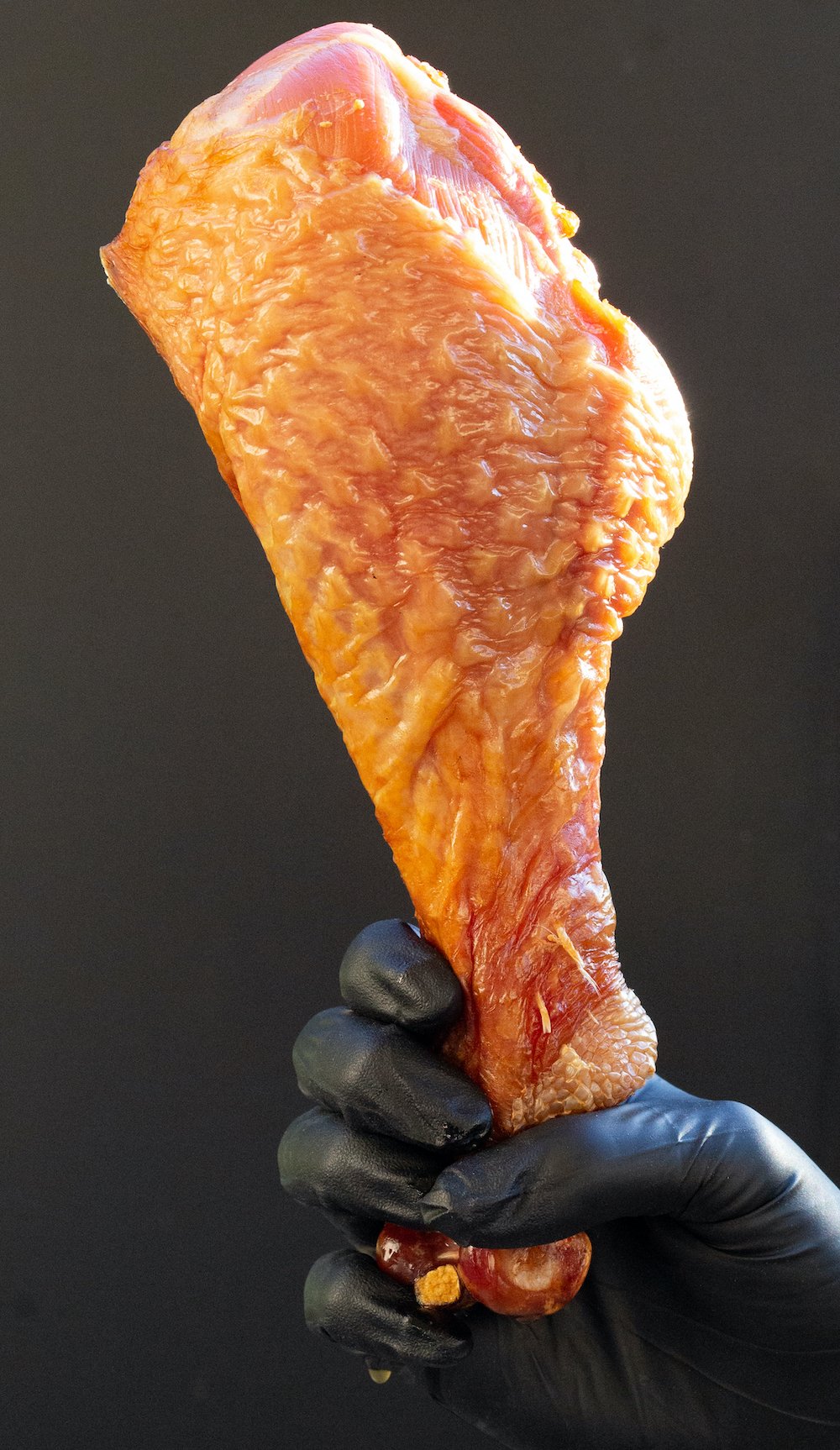 A hand holds a very large smoked turkey leg in front of a black electric smoker