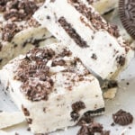 Two pieces of cookies and cream fudge sit on a white background surrounded by Oreos and cookie crumbs