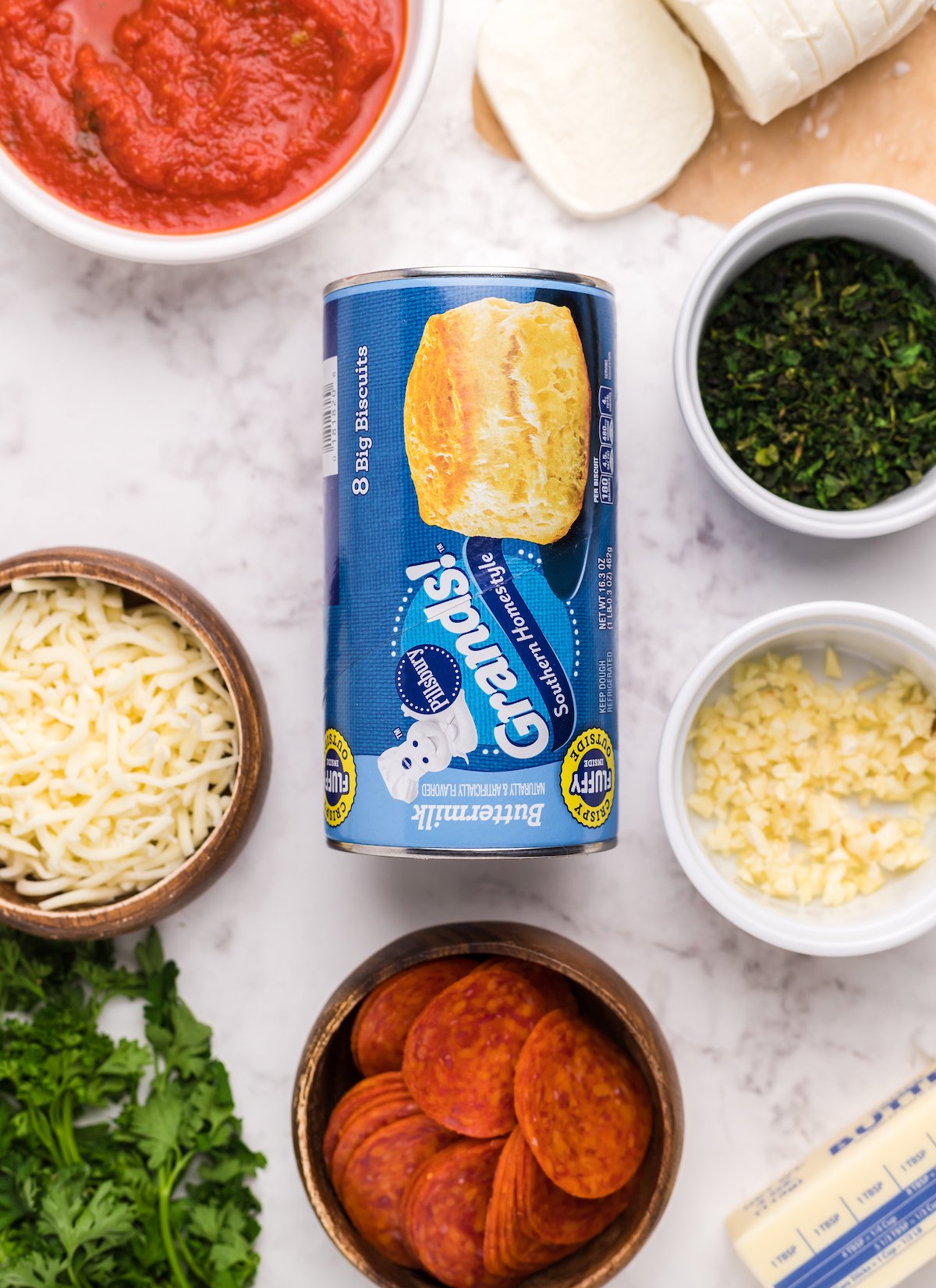 Overhead look at the ingredients to make biscuit pizza casserole: canned Grands! Biscuits, shredded mozzarella, fresh mozzarella, pepperonis, butter, minced garlic, and parsley. 