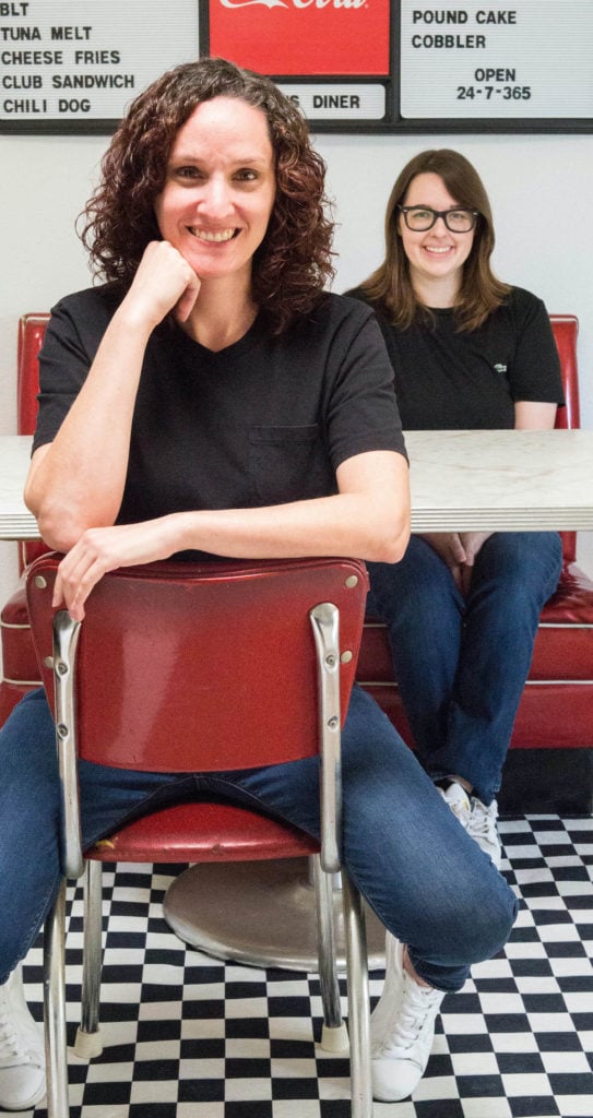 Two women sit at a diner table with a red booth, red chair, and black and white checkered floor