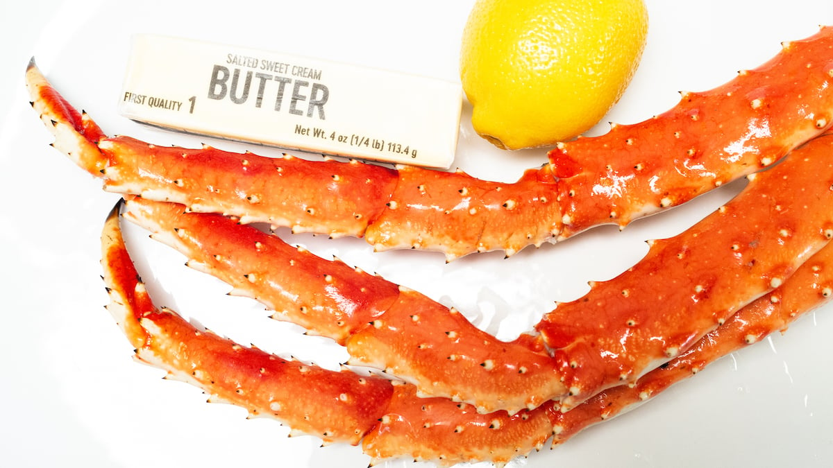 A cluster of king crab legs sit on a white platter next to a stick of butter and a lemon