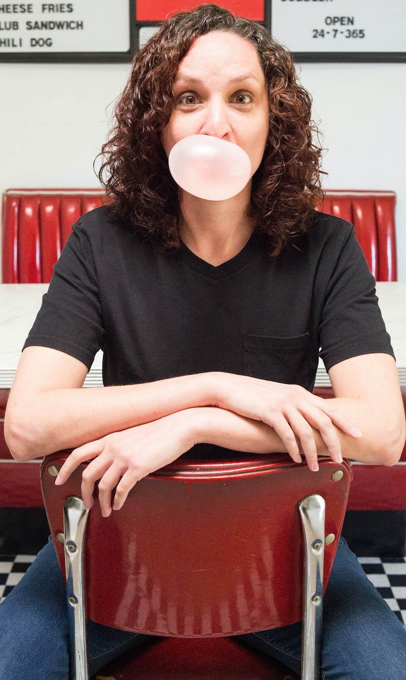 A women in a black t-shirt sits at a red diner chair and blows a bubble of pink bubblegum