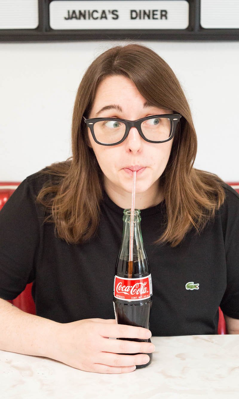 A woman in a black t-shirt sips on a coke in a bottle while sitting at a diner table