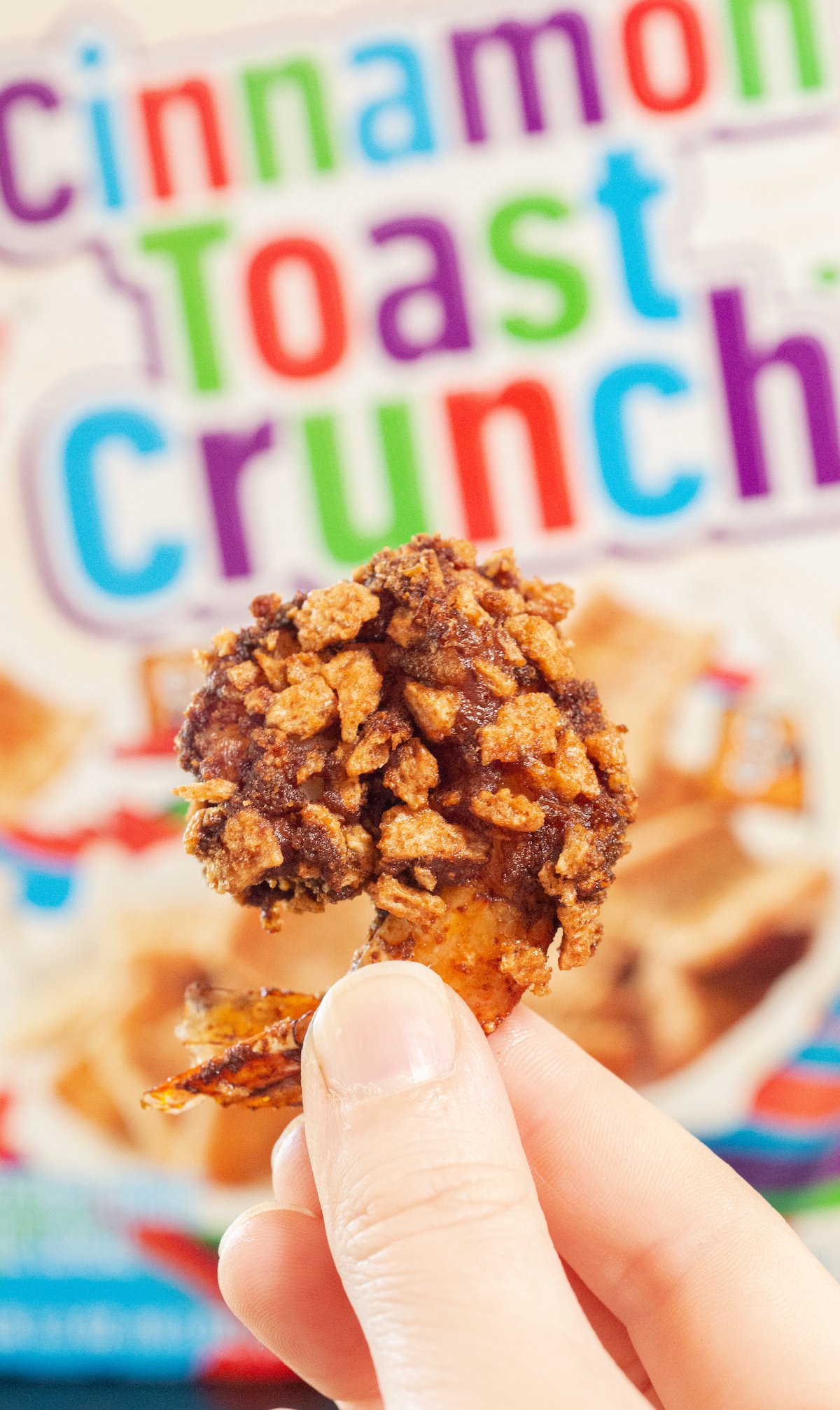 A finger holds up a piece of fried shrimp that's been breaded with cinnamon toast crunch in front of the cereal box.