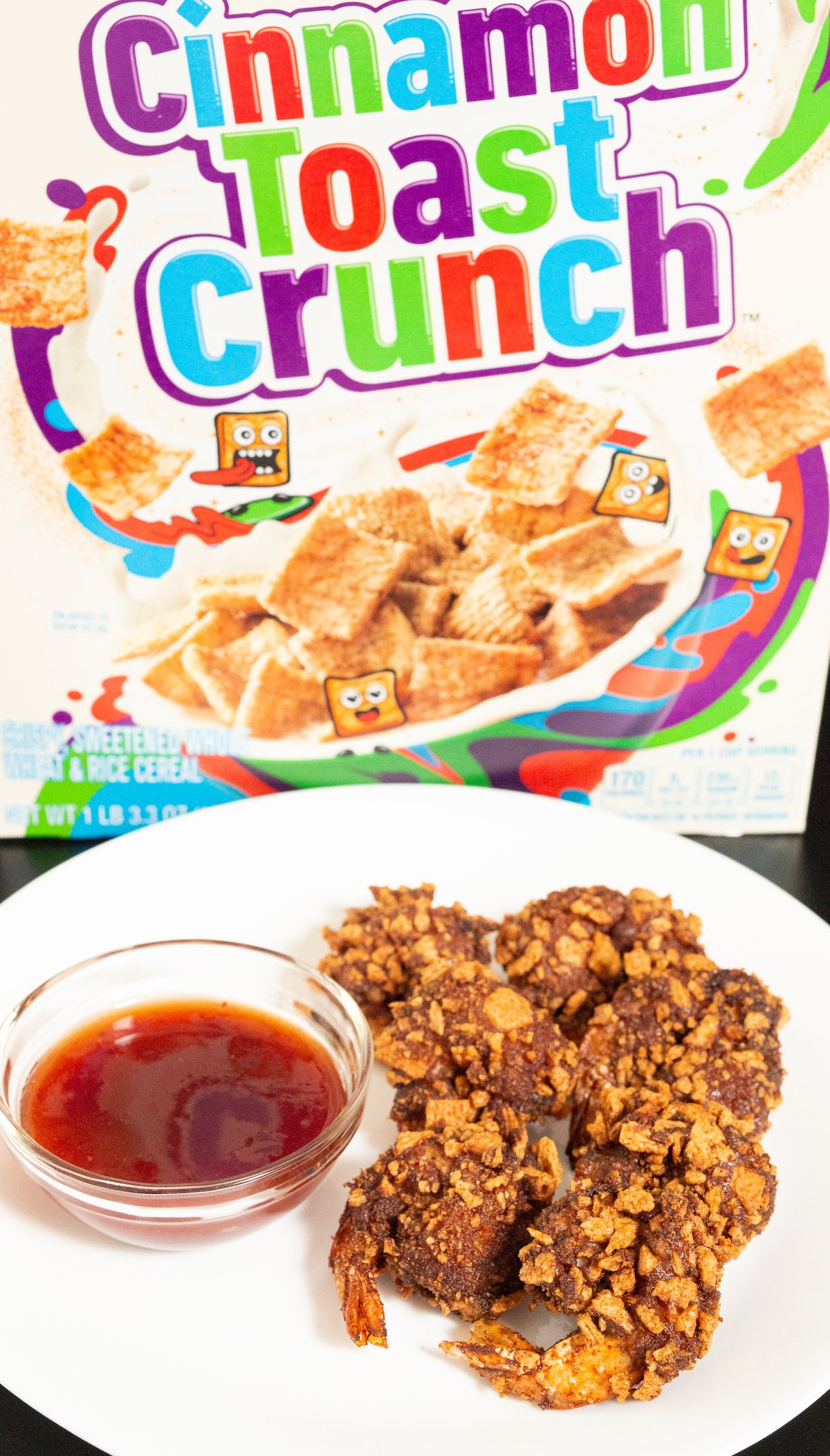 Six Cinnamon Toast Crunch battered and fried shrimp sit on a white plate next to strawberry habanero dipping sauce and in front of a Cinnamon Toast Crunch cereal box.