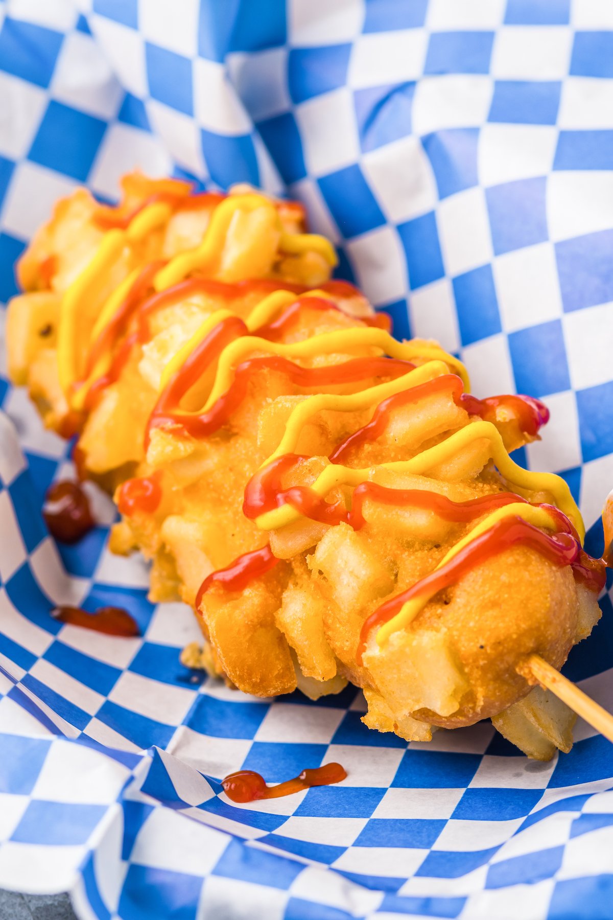 A Korean french fry coated corn dog drizzled with ketchup and mustard sits in a food basket with blue checkered parchment paper.