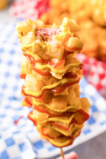 A corn dog that's covered in a french fry batter and drizzled with mustard and ketchup has a bite taken out of it.