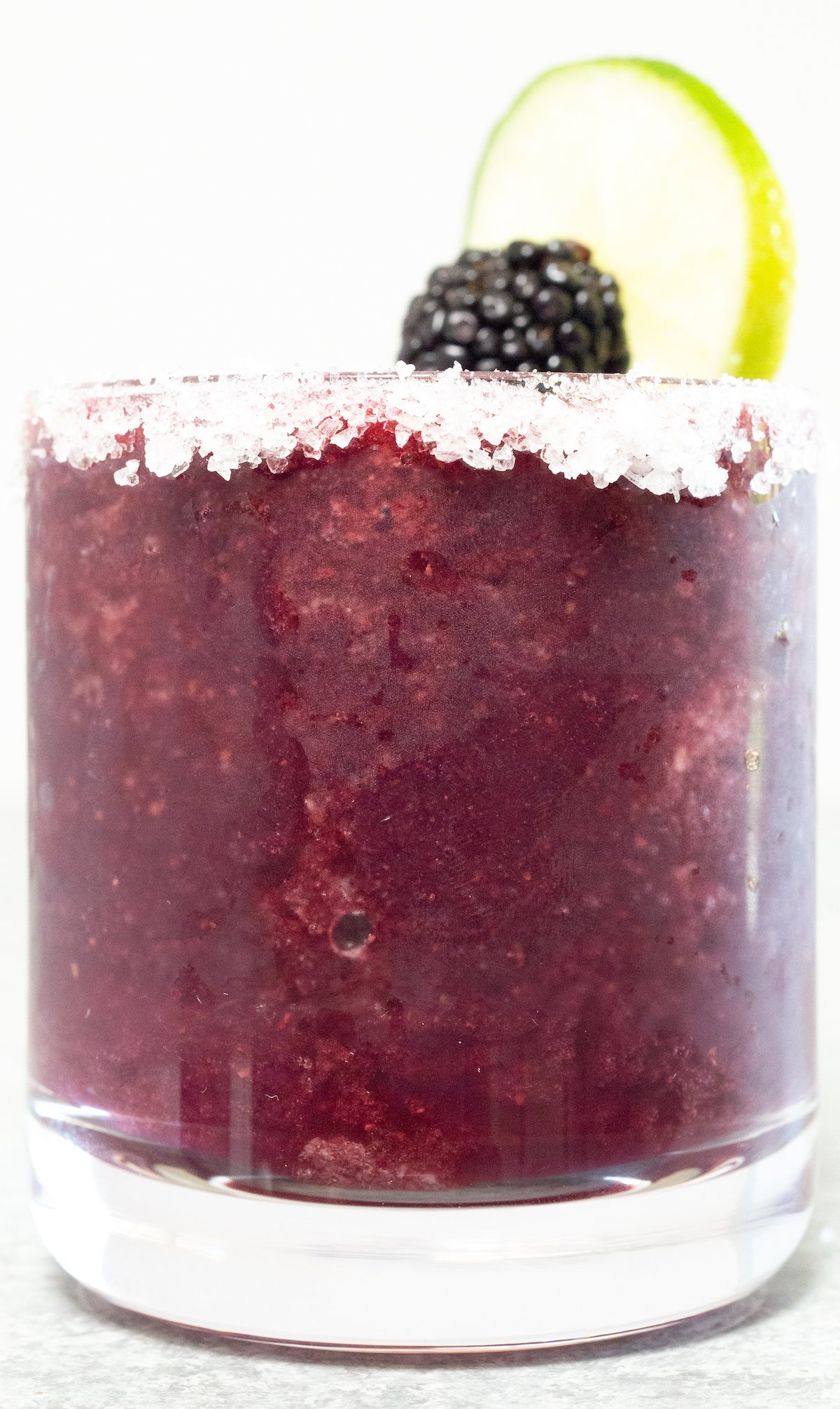 A glass filled with a purple blackberry margarita sits on a white background.