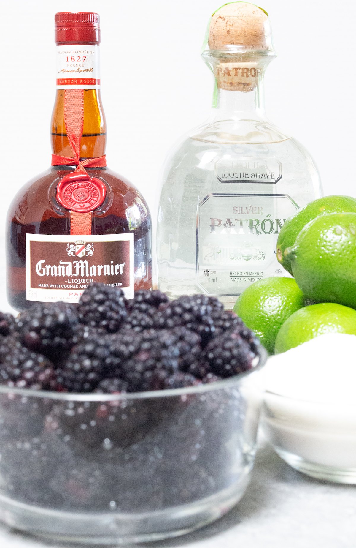 The ingredients for blackberry margaritas on a white background. Grand marnier, Patron, blackberries, limes, and sugar.