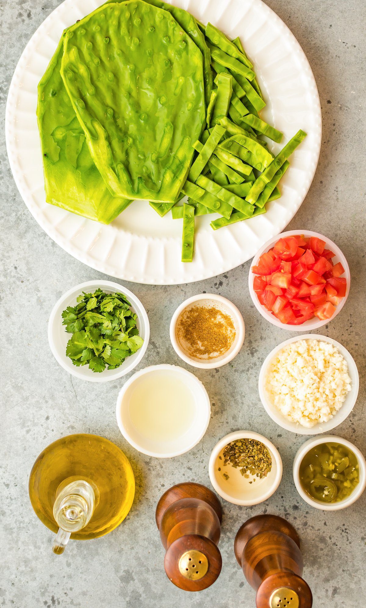 Overhead view of the ingredients for cactus salad arranged in white prep bowls: fresh cactus paddles, cilantro, cumin, chopped tomatoes, queso fresco, oregano, pickled jalapenos, salt, pepper, and olive oil.