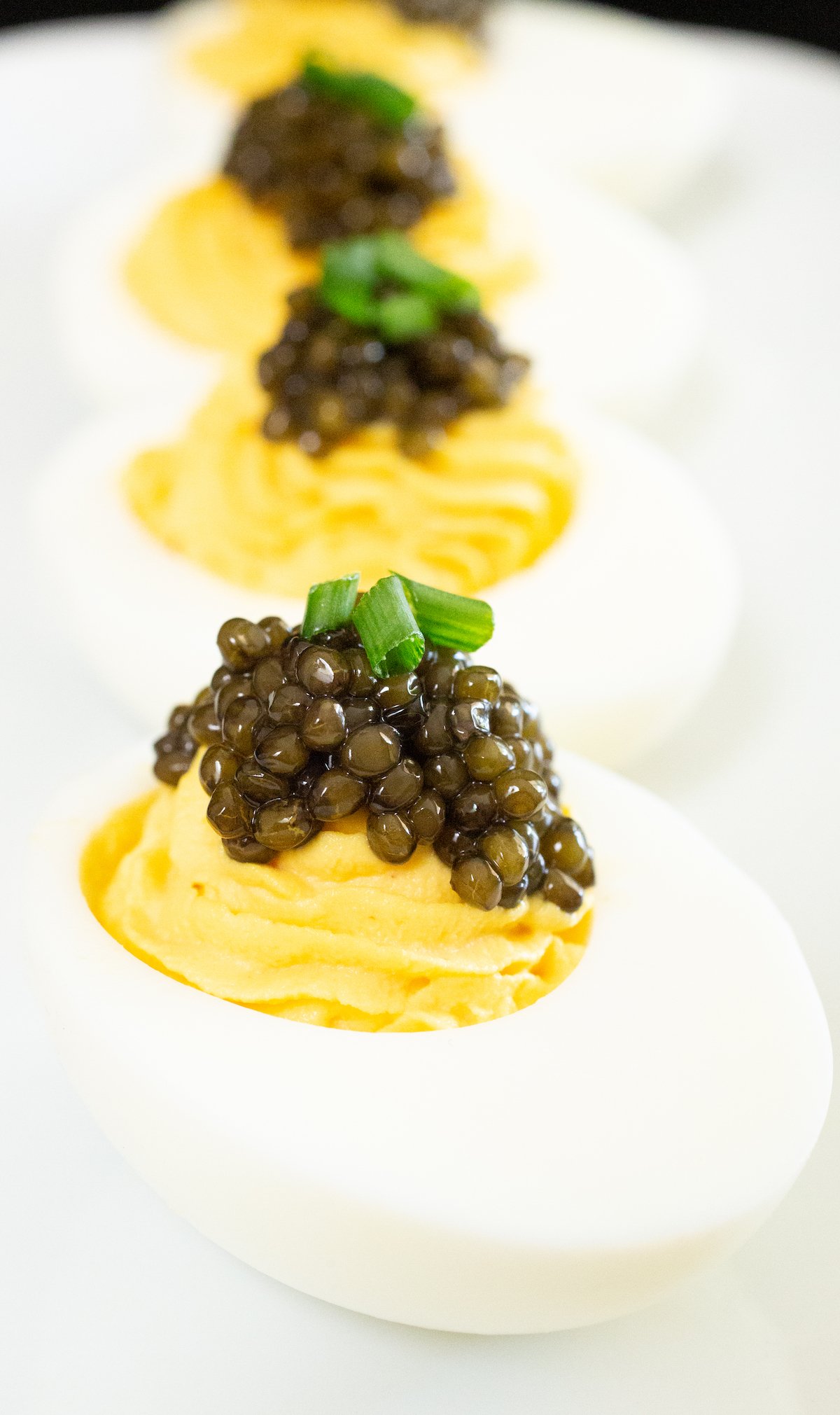 Four deviled eggs topped with caviar and chives sit in a row on a white plate.