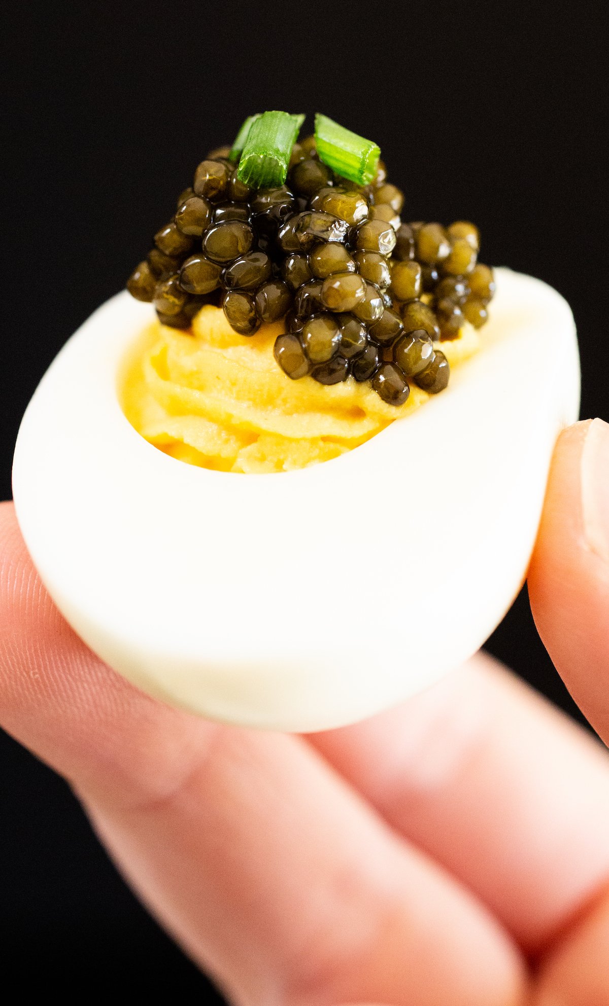 A hand holds up a deviled egg topped with caviar against a black background
