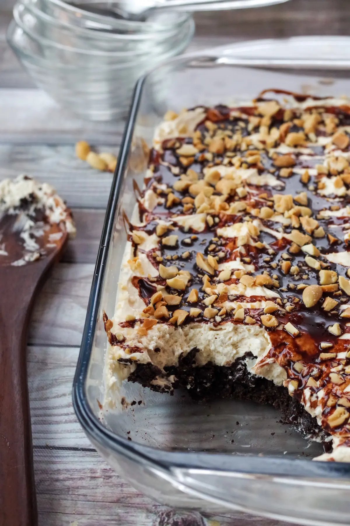 A glass casserole dish is filled with a drumstick dessert that has a layer of chocolate cake, then whipped topping that's drizzled with chocolate sauce and garnished with crushed peanuts.