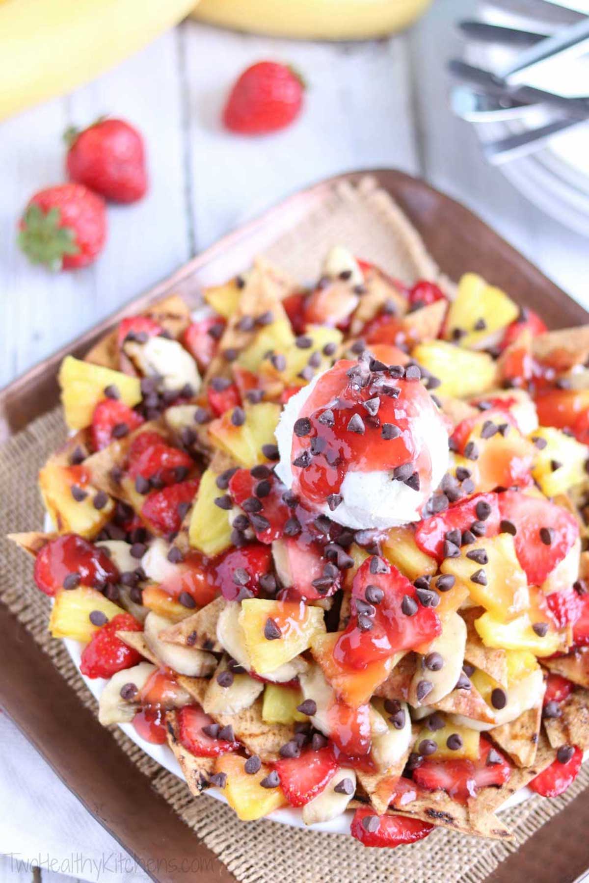 Overhead view of a tray of dessert nachos covered in grilled strawberries, grilled bananas, and chocolate chips - then topped with a scoop of vanilla ice cream.